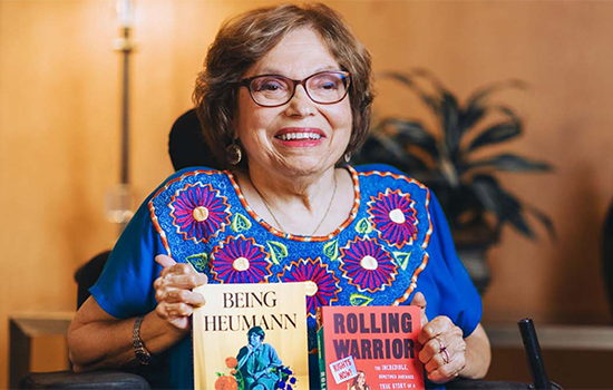 Activist Judy Heumann holding her two books, Being Heumann: An Unrepentant Memoir of a Disability Rights Activist and Rolling Warrior.